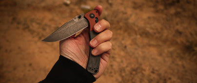 Hand grips folding knife with textured drop-point blade and red handle, set against earthy outdoor background.