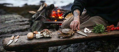 Tattooed hands prep a crab and mushrooms on a rustic wooden plank by a campfire, showcasing sharp outdoor kitchen knives.