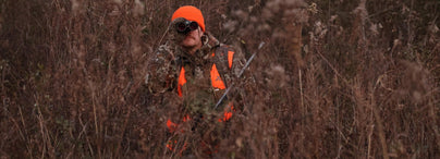 Hunter in orange gear and camo peers through binoculars into tall, dry brush during a quiet whitetail hunt.