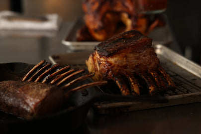 Glistening glazed pork rack roast with charred bones resting on a metal tray in warm, moody kitchen lighting.