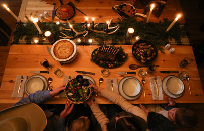 Hands share a salad bowl over a candlelit wooden table set with cornbread, roasted potatoes, and lamb chops.