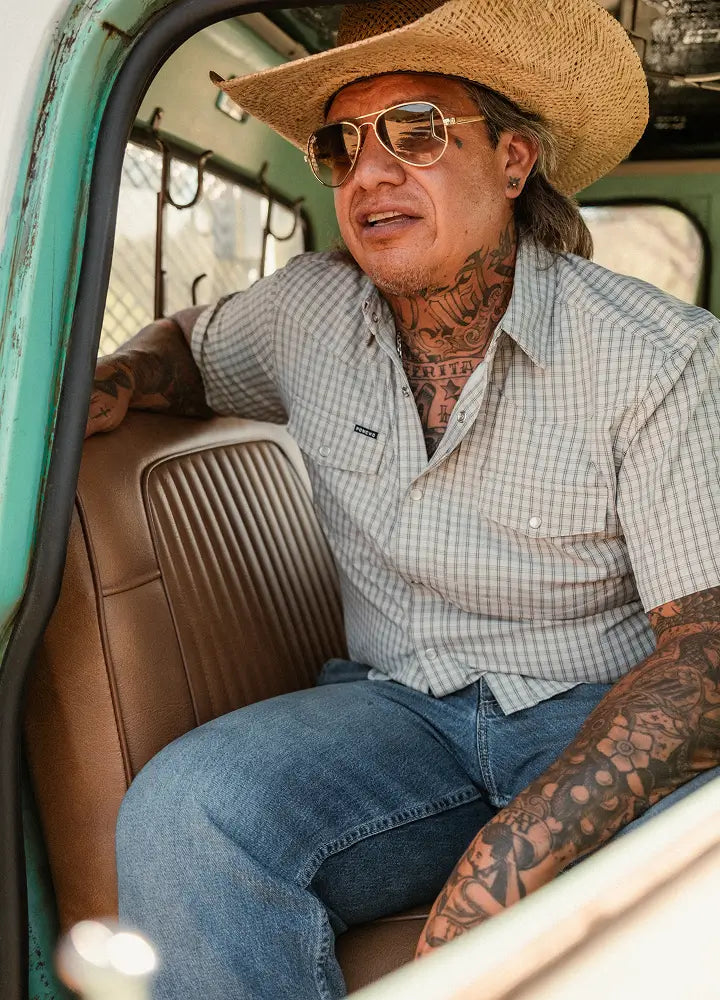Tattooed man in straw cowboy hat and aviator sunglasses sits inside vintage teal truck with brown leather seat.