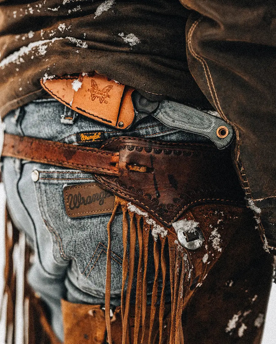 Close-up of a person in Wrangler jeans and fringed chaps, with a Benchmade knife tucked into their belt.