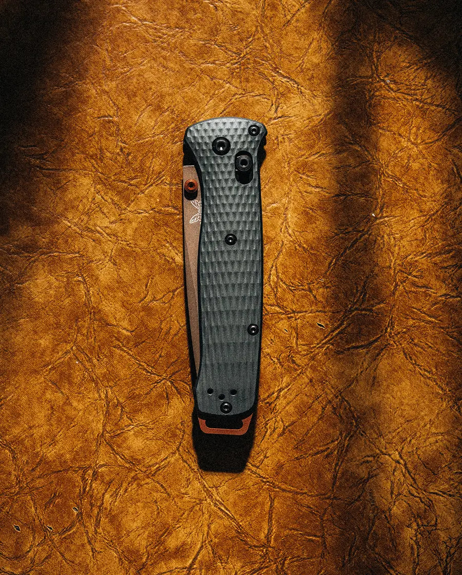 Closed folding knife with textured gray handle and copper accents rests on wrinkled brown leather under warm lighting.